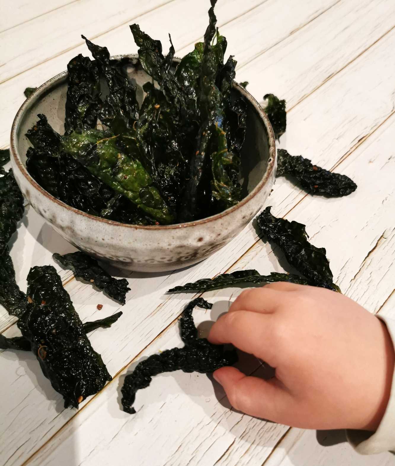 Skål med grönkålschips på ett bord, med en hand som sträcker sig mot chipsen.