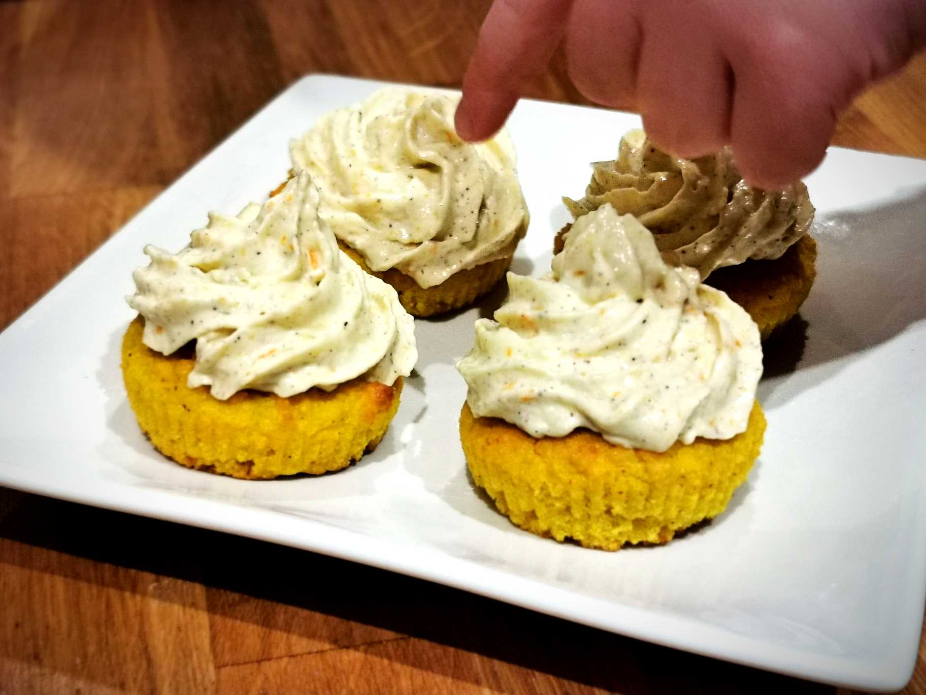 Fyra cupcakes med gräddfrosting på en vit tallrik, en hand pekar på en av dem.