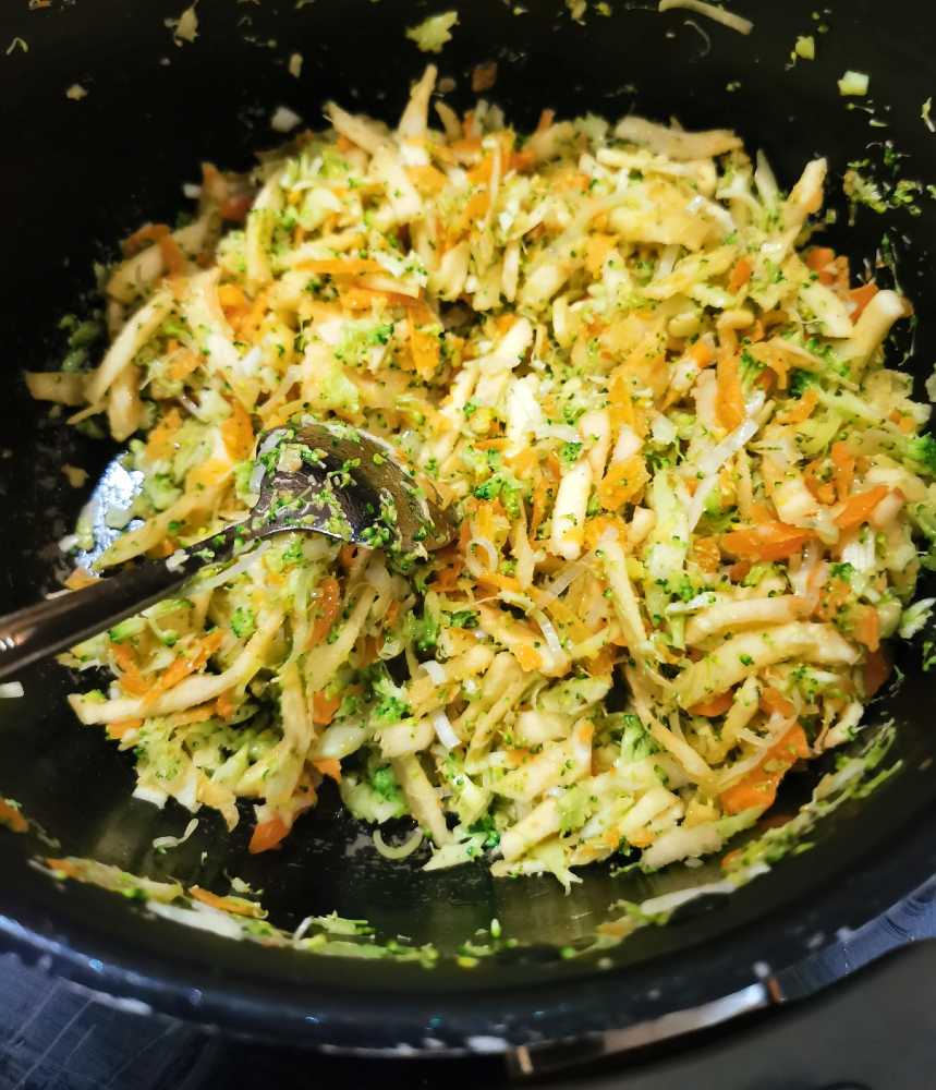Blandning av riven kål, äpplen, morötter och broccoli i en skål med en sked.