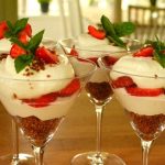 Fyra glas med jordgubbsdessert, grädde och mynta på toppen, placerade på ett träbord.