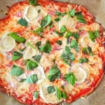 En rund pizza med ost, skinka, lök och färska örter på en bakplåt.