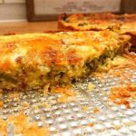 En skiva grönsaksquiche med ost på en metallplåt, med en annan quiche i bakgrunden.