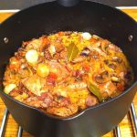 En svart gryta med kött, svamp, grönsaker och kryddor på en spis.