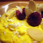 Gul dessert toppad med mandelspån och frysta hallon på en tallrik.