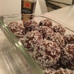 En skål med chokladbollar täckta av kokosflingor och en flaska ingefäraextrakt i bakgrunden.