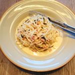 Tallrik med pasta, bacon och grönsaker, serverad med en gaffel och sked.