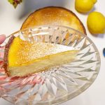 En bit citroncheesecake på ett glasfat med en hel cheesecake och citroner i bakgrunden.