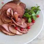 Skivad rökt köttbit på en tallrik med lingon och färska örter.