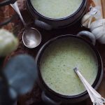 Två skålar med broccolisoppa, en sked ligger bredvid. Vitlök syns i bakgrunden.