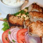 Grillade köttspett serverade med grönsaker, tomater och en skål med sås.