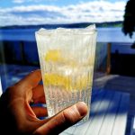 Ett glas med kolsyrad dryck och isbitar hålls i handen framför en sjöutsikt.