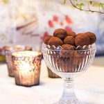 En glasbehållare med chokladtryfflar täckta av kakao, placerad på ett bord med ljus i bakgrunden.